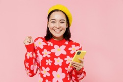 Happy woman of Asian ethnicity 20s wear red knitted sweater beret hold in hand use mobile cell phone do winner gesture isolated on plain pastel pink background studio. People lifestyle fashion concept