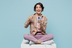 Full body young smiling cheerful happy woman 20s in brown shirt sit on white chair use point finger on mobile cell phone isolated on pastel plain light blue background studio People lifestyle concept