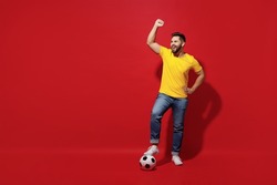 Full size body length young bearded man football fan in yellow t-shirt cheer up support favorite team hold foot on soccer ball do winner gesture isolated on plain dark red background studio portrait