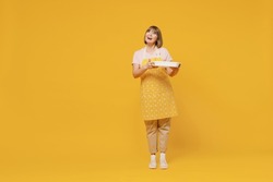 Full size elderly smiling happy fun housekeeper housewife woman 50s in orange apron hold casserole dinner meal look aside isolated plain on yellow background studio. People household lifestyle concept