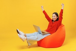 Full size body length young woman of Asian ethnicity 20s in casual clothes sit in bag chair hold use work on laptop pc computer doing winner gesture isolated on plain yellow background studio portrait