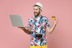 Funny traveler tourist man in summer clothes hat using laptop pc computer booking hotel hold credit bank card isolated on pink background. Passenger traveling on weekends. Air flight journey concept