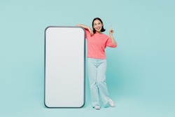 Full body young woman of Asian ethnicity in pink sweater stand near big mobile cell phone with blank screen workspace area point finger up with new idea isolated on pastel plain light blue background