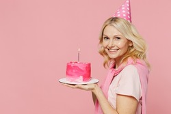 Side profile view elderly smiling cool happy woman 50s wear t-shirt birthday hat hold cake with candle look camera isolated on plain pastel pink background studio Celebration party holiday concept.