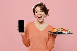 Young surprised woman in sweater hold makizushi sushi roll served on black plate traditional japanese food use mobile cell phone blank screen workspace area isolated on plain pastel pink background.