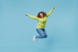 Young overjoyed excited chubby overweight plus size big fat fit woman wear green sweater jump high with outstretched hands isolated on plain blue background studio portrait. People lifestyle concept