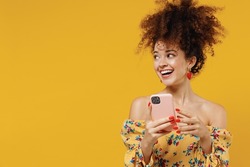 Young happy smiling woman 20s with culry hair in casual clothes use hold mobile cell phone look aside on copy space workspace area isolated on plain yellow background studio. People lifestyle concept