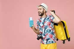 Funny young traveler tourist man in summer clothes hat photo camera hold suitcase passport ticket looking aside isolated on pink background. Passenger traveling on weekend. Air flight journey concept