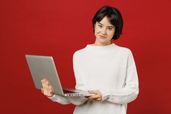 Young smiling happy fun woman 20s wear white knitted sweater hold use work on laptop pc computer chatting online surfing internet isolated on plain red color background studio People lifestyle concept