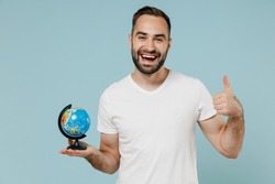 Young smiling happy fun geography teacher man wear blank print design white t-shirt hold in hands Earth world globe show thumb up gesture isolated on plain pastel light blue color background studio.