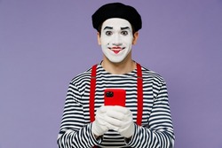 Smiling fun amazing marvelous ecstatic young mime man with white face mask wears striped shirt beret hold in hand use mobile cell phone isolated on plain pastel light violet background studio portrait