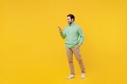 Full body side view smiling happy young man 20s wearing mint knitted sweater hold in hand use mobile cell phone isolated on plain yellow background studio portrait. People lifestyle fashion concept