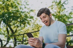 Overjoyed exultant young happy man 20s in blue t-shirt sit on bench using mobile cell phone chat online scream rest relax in sunshine spring green city park outdoor on nature Urban leisure concept.
