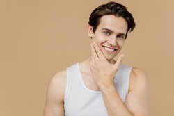 Attractive smiling calm young man he 20s perfect skin wearing bundershirt touch face after shaving isolated on light pastel beige background studio. Skin care healthcare cosmetic procedures concept.