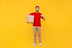 Full body smiling fun happy delivery guy employee man in red cap T-shirt uniform workwear work as dealer courier hold cardboard box show thumb up isolated on plain yellow background studio portrait.