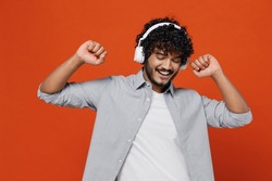 Vivid fun cheerful young bearded Indian man 20s years old wears blue shirt listen music in headphones dance fooling around gesticulating hands enjoy isolated on plain orange background studio portrait