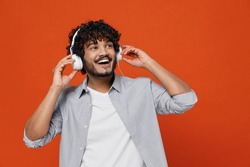 Cheerful exultant happy young bearded Indian man 20s years old wears blue shirt listen music in headphones dance have fun gesticulating hands relax isolated on plain orange background studio portrait