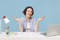 Young employee business woman in casual shirt set microphone headset for helpline assistance sit work at office desk with pc laptop spreading hands isolated on pastel blue background studio portrait