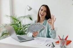Young employee business woman in blue shirt hold fan cash money, dollar banknotes show ok gesture sit work at workplace white desk with laptop pc computer at office indoors. Achievement career concept