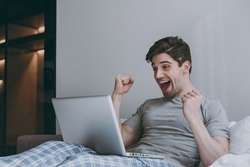 Lucky overjoyed excited young man in pajamas grey t-shirt sit in bed hold use work on laptop pc computer clench fist rest relax at home indoors bedroom Good mood sleeping night morning bedtime concept