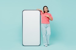 Full body young smiling happy woman of Asian ethnicity 20s in pink sweater point finger on big mobile cell phone with blank screen workspace area isolated on pastel plain light blue background studio