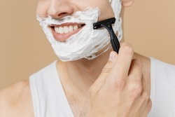 Cropped smiling young man he 20s perfect skin in undershirt face covered with foam shaving with blade isolated on light pastel beige background studio Skin care healthcare cosmetic procedures concept