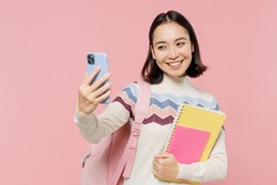 Teen student girl of Asian ethnicity in sweater backpack hold books doing selfie shot on mobile cell phone isolated on pastel plain light pink color background Education in university college concept