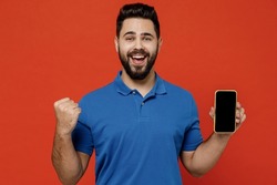 Young smiling fun happy successful cheerful man 20s wear basic blue t-shirt hold in hand use mobile cell phone with blank screen workspace area do winner gesture isolated on plain orange background.