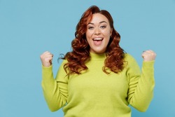 Young excited fun overjoyed chubby overweight plus size big fat fit woman wear green sweater do winner gesture clench fist isolated on plain blue background studio portrait. People lifestyle concept.