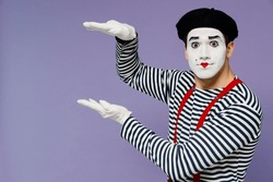 Charismatic amazing ecstatic young mime man with white face mask wears striped shirt beret hands aside like holding carrying something isolated on plain pastel light violet background studio portrait