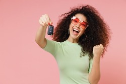Exultant happy excited young curly latin woman 20s wears mint t-shirt sunglasses hold vehicle key doing winner gesture isolated on plain pastel light pink background studio portrait. Car sales concept