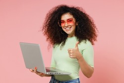 Fun young curly latin woman 20s years old wears mint t-shirt sunglasses hold use work on laptop pc computer showing thumb up like gesture isolated on plain pastel light pink background studio portrait