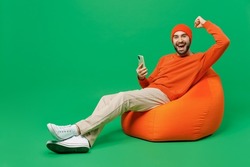Full body young fun happy man 20s wear orange sweatshirt hat sit in bag chair hold in hand use mobile cell phone do winner gesture isolated on plain green background studio. People lifestyle concept
