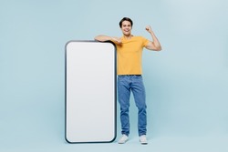 Full body young man in yellow t-shirt stand near big mobile cell phone with blank screen workspace area do winner gesture isolated on plain pastel light blue background studio People lifestyle concept