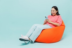 Full body young smiling happy woman of Asian ethnicity 20s wearing pink sweater sit in bag chair hold in hand use mobile cell phone isolated on pastel plain light blue color background studio portrait