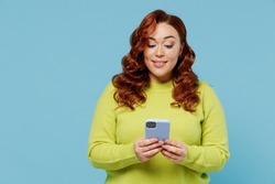 Young smiling cheerful happy fun chubby overweight plus size big fat fit woman in green sweater hold use mobile cell phone isolated on plain blue background studio portrait. People lifestyle concept