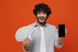 Jubilant exultant young bearded Indian man 20s years old wears blue shirt using mobile cell phone hold in hand fan of cash money in dollar banknotes isolated on plain orange background studio portrait