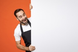 Young excited man barista bartender barman employee in apron white t-shirt work coffee shop big white blank billboard for promo content text image place isolated on orange background Business startup