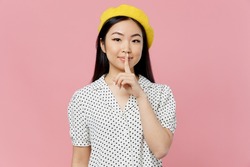 Young secret woman of Asian ethnicity 20s in white polka dot t-shirt yellow beret say hush be quiet with finger on lips shhh gesture isolated on plain pastel pink background. People lifestyle concept