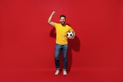 Full size body length happy young bearded man football fan in yellow t-shirt cheer up support favorite team hold soccer ball doing winner gesture isolated on plain dark red background studio portrait