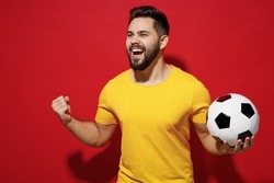 Jubilant fun young bearded man football fan in yellow t-shirt cheer up support favorite team hold soccer ball look aside clenching fists say yes isolated on plain dark red background studio portrait