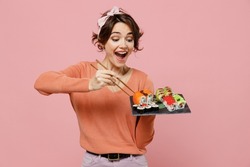 Young excited amazed cool fun woman 20s in casual clothes hold in hand eat makizushi sushi roll served on black plate traditional japanese food chopsticks isolated on plain pastel pink background.