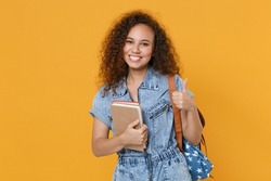 Smiling young african american girl student in denim clothes, backpack isolated on yellow background studio portrait. Education in high school university college concept. Hold books, showing thumb up