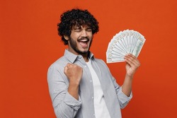 Young bearded Indian man 20s years old wears blue shirt holding fan of cash money in dollar banknotes doing winner gesture celebrate clenching fists isolated on plain orange background studio portrait