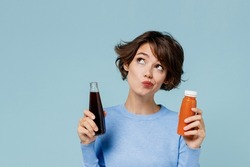 Young minded woman 20s wear casual sweater hold soda cola water vegetable smoothie choose between junk food and healthy isolated on plain pastel light blue background. People lifestyle food concept
