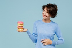 Young smiling cool woman 20s in casual sweater look camera hold donuts dessert put hand on belly isolated on plain pastel light blue background studio. People lifestyle junk unhealthy food concept