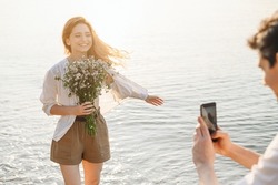 Sunlit young couple family man woman in white clothes hold flowers bouquet take photo on mobile cell phone rest together at sunrise over sea beach ocean outdoor seaside in summer day sunset evening.
