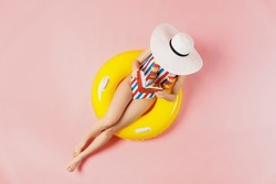 Top view full body young woman in striped swimsuit lies on inflatable rubber ring pool read book cover face with hat isolated on plain pastel pink background. Summer vacation sea rest sun tan concept