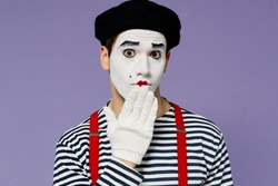 Close up confused preoccupied young mime man with white face mask wears striped shirt beret cover mouth with hand doing oops gesture isolated on plain pastel light violet background studio portrait