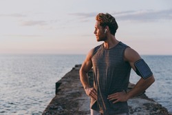 Young strong sporty athletic toned fit sportsman man in sports clothes earphones warm up train with mobile phone akimbo at sunrise sun dawn over sea beach outdoor on pier seaside in summer day morning